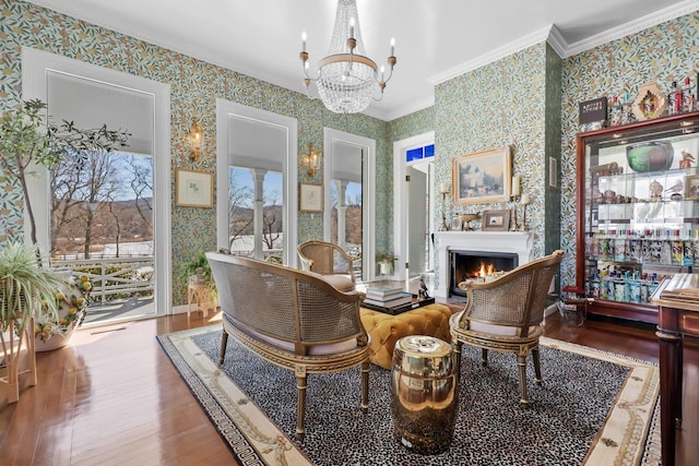 sitting room with a warm lit fireplace, ornamental molding, wood finished floors, and wallpapered walls