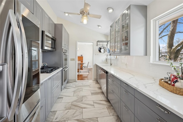 kitchen with marble finish floor, appliances with stainless steel finishes, gray cabinets, light stone countertops, and glass insert cabinets