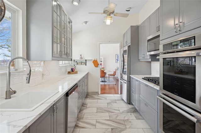 kitchen featuring gray cabinets, glass insert cabinets, appliances with stainless steel finishes, a sink, and light stone countertops