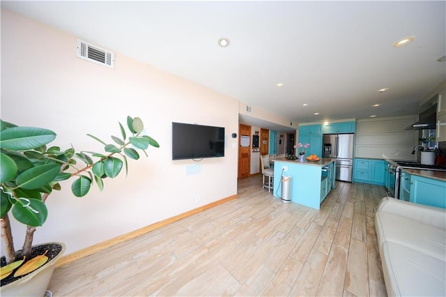 kitchen with wall chimney exhaust hood, blue cabinets, stainless steel appliances, and light hardwood / wood-style flooring