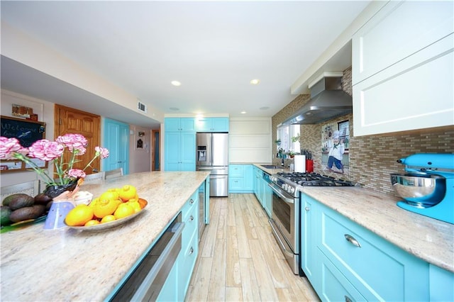 kitchen with blue cabinetry, appliances with stainless steel finishes, light hardwood / wood-style floors, decorative backsplash, and wall chimney exhaust hood