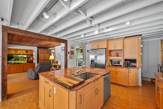 kitchen featuring a kitchen island with sink, stainless steel appliances, a sink, dark stone counters, and beamed ceiling