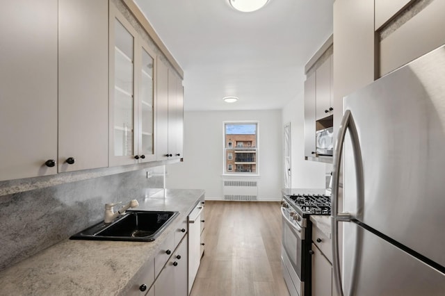 kitchen featuring radiator, appliances with stainless steel finishes, light stone counters, glass insert cabinets, and a sink