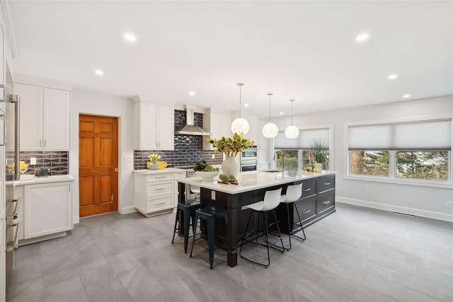 kitchen with decorative light fixtures, light countertops, a kitchen island with sink, white cabinetry, and wall chimney range hood