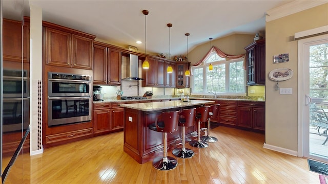 kitchen with wall chimney exhaust hood, light wood-style flooring, appliances with stainless steel finishes, a kitchen island, and a kitchen breakfast bar