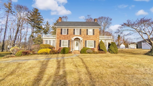 colonial inspired home featuring brick siding, a chimney, a front yard, and a balcony