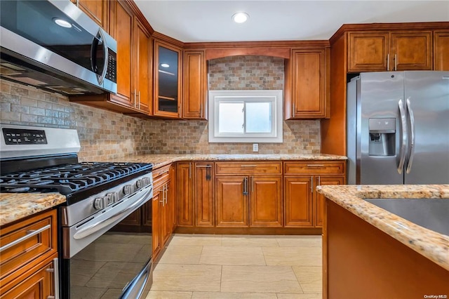 kitchen featuring appliances with stainless steel finishes, brown cabinetry, glass insert cabinets, and light stone countertops