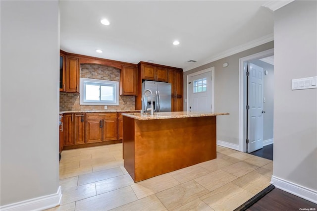 kitchen with light stone counters, a center island with sink, backsplash, brown cabinetry, and stainless steel fridge with ice dispenser