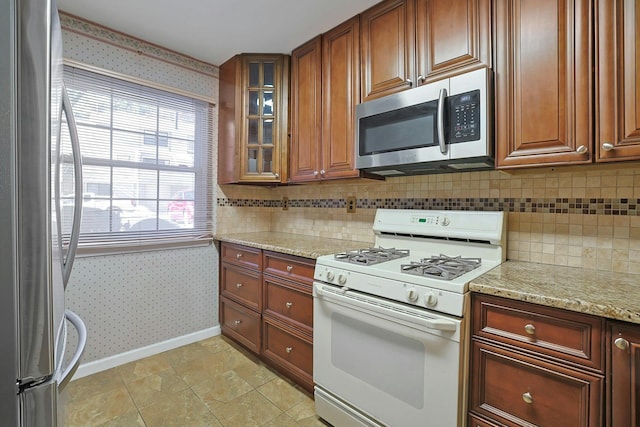 kitchen with wallpapered walls, glass insert cabinets, baseboards, light stone counters, and stainless steel appliances