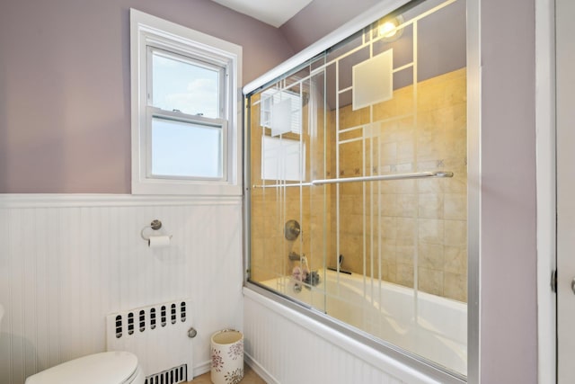 full bathroom featuring bath / shower combo with glass door, radiator heating unit, wainscoting, and toilet