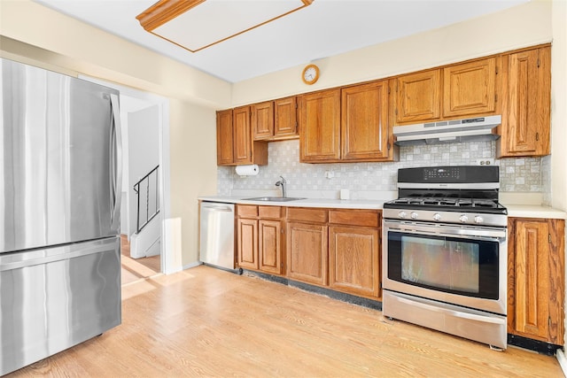 kitchen with brown cabinets, stainless steel appliances, light countertops, under cabinet range hood, and a sink