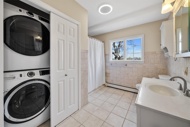 washroom with stacked washer and dryer, a sink, a baseboard heating unit, light tile patterned flooring, and tile walls