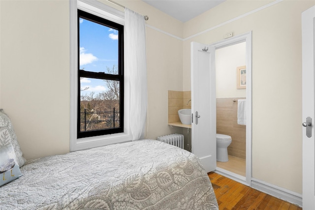 bedroom with radiator, a wainscoted wall, wood finished floors, ensuite bathroom, and tile walls