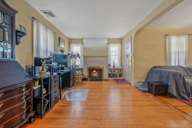 home office with baseboard heating, a brick fireplace, visible vents, and light wood finished floors