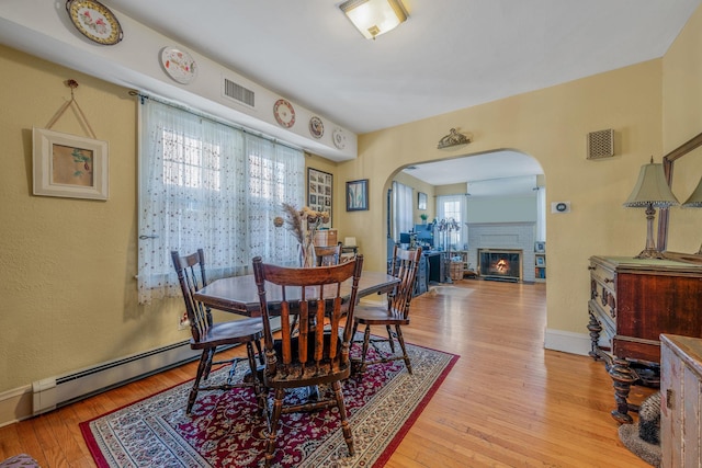 dining area with visible vents, arched walkways, a glass covered fireplace, baseboard heating, and light wood-style floors