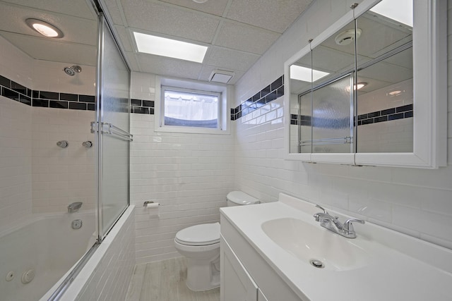 full bath featuring a drop ceiling, toilet, shower / bath combination with glass door, vanity, and tile walls