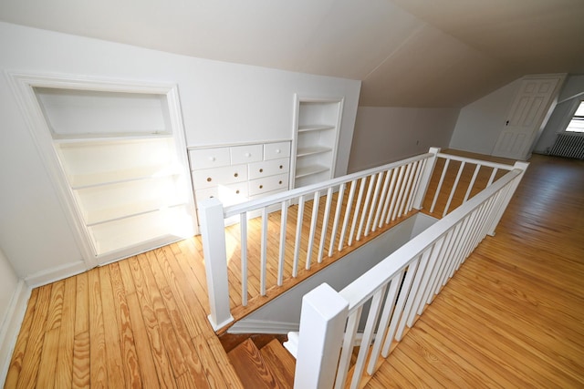 corridor with built in features, vaulted ceiling, wood finished floors, and an upstairs landing