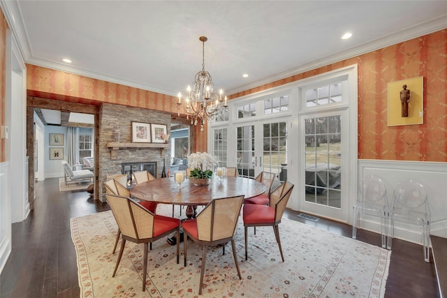 dining room featuring wallpapered walls, ornamental molding, a stone fireplace, wainscoting, and french doors