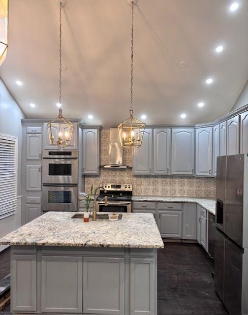 kitchen featuring a kitchen island with sink, appliances with stainless steel finishes, wall chimney range hood, gray cabinets, and decorative light fixtures