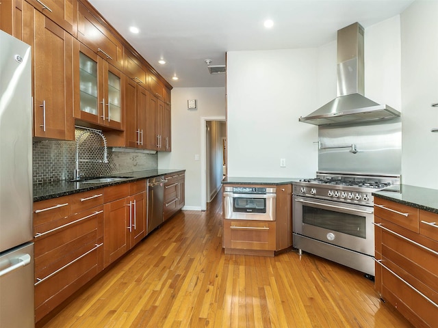 kitchen featuring tasteful backsplash, light wood-style flooring, appliances with stainless steel finishes, brown cabinetry, and wall chimney range hood