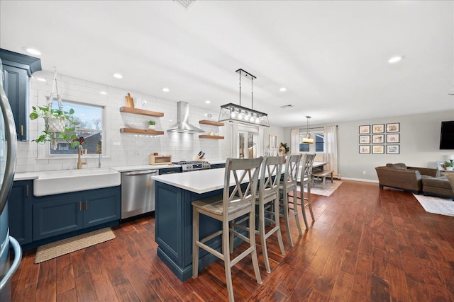 kitchen with a sink, blue cabinetry, stainless steel dishwasher, wall chimney exhaust hood, and dark wood finished floors