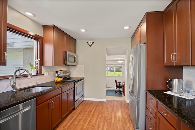 kitchen with a baseboard radiator, appliances with stainless steel finishes, a sink, dark stone counters, and light wood-type flooring