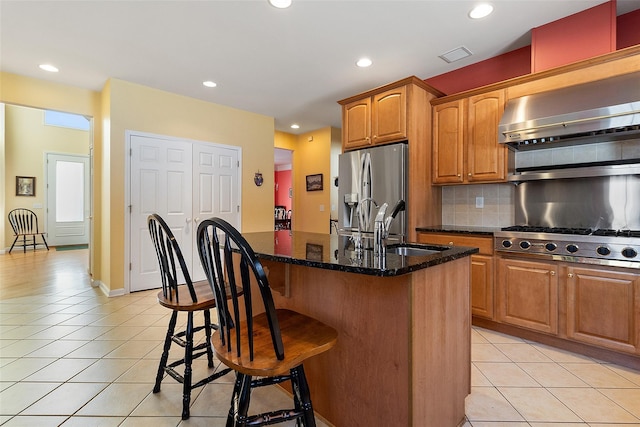 kitchen featuring appliances with stainless steel finishes, light tile patterned flooring, a center island with sink, and dark stone countertops