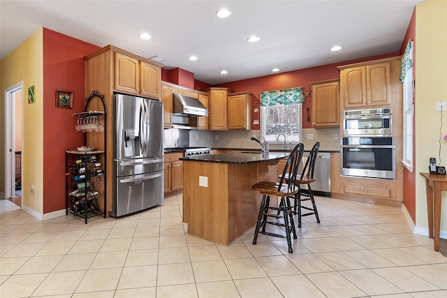 kitchen featuring appliances with stainless steel finishes, wall chimney range hood, an island with sink, light tile patterned floors, and decorative backsplash
