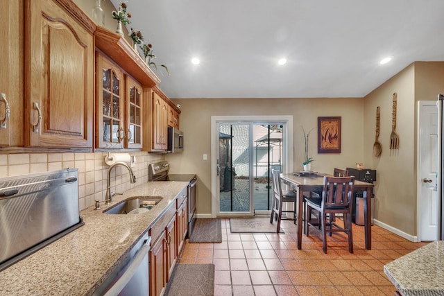 kitchen featuring brown cabinetry, glass insert cabinets, light stone countertops, stainless steel appliances, and a sink