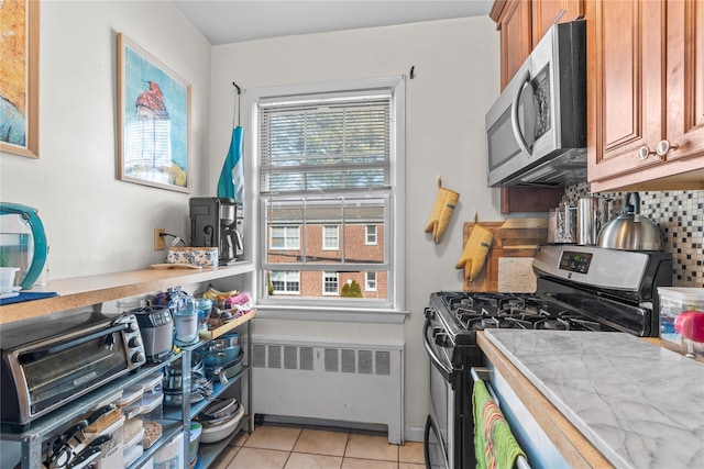 kitchen with light tile patterned floors, appliances with stainless steel finishes, radiator, tasteful backsplash, and brown cabinetry