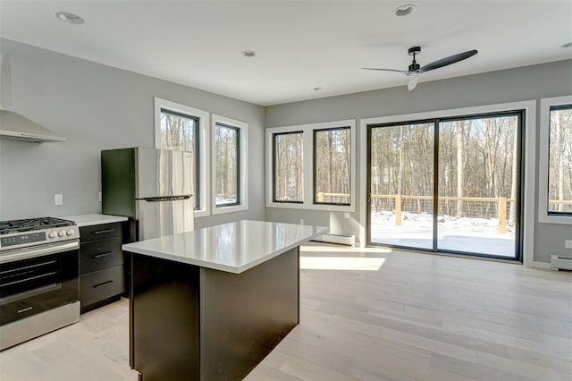 kitchen with appliances with stainless steel finishes, a center island, light wood-type flooring, a baseboard heating unit, and wall chimney range hood