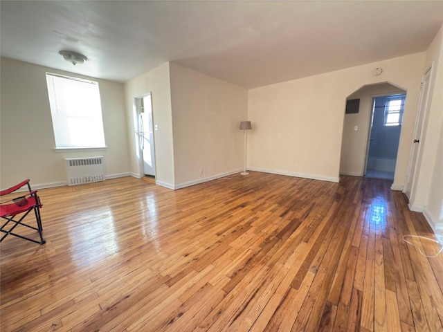 empty room with arched walkways, radiator heating unit, light wood-style flooring, and baseboards