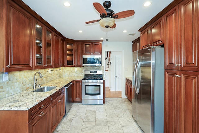 kitchen with a sink, light stone counters, stainless steel appliances, glass insert cabinets, and ceiling fan