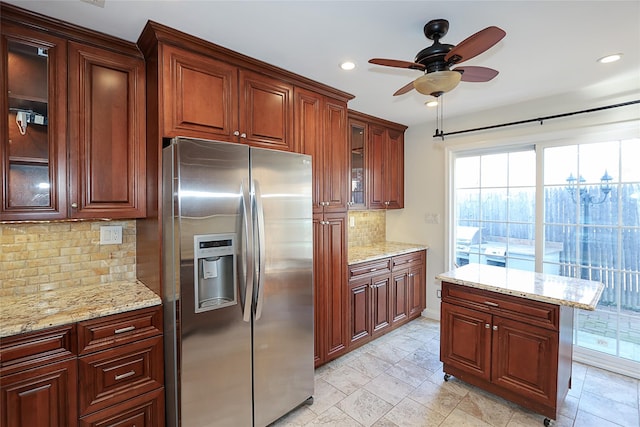 kitchen featuring glass insert cabinets, ceiling fan, light stone counters, decorative backsplash, and stainless steel fridge