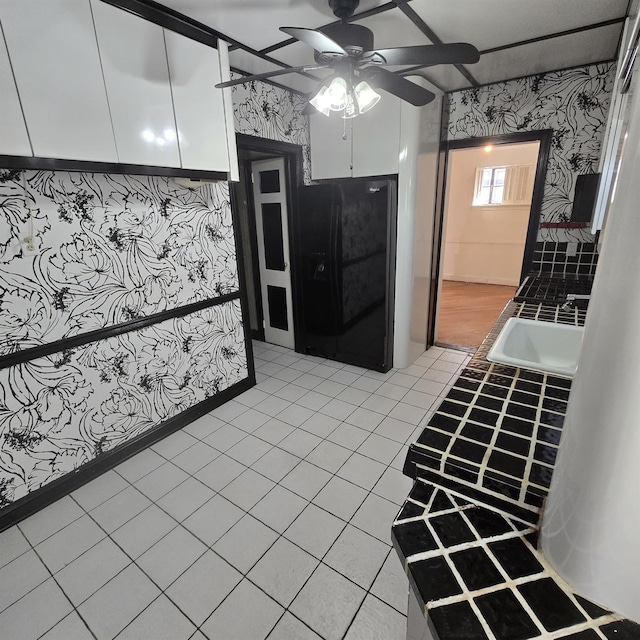 kitchen featuring light tile patterned floors, ceiling fan, white cabinetry, black fridge, and wallpapered walls