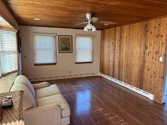 unfurnished living room with dark wood finished floors, a baseboard heating unit, a ceiling fan, wood ceiling, and wood walls