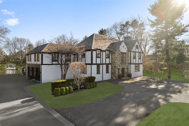 english style home featuring driveway, stone siding, stucco siding, a chimney, and a front yard