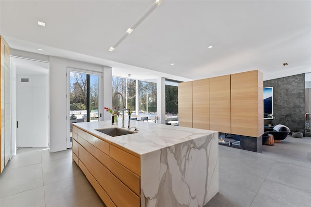 kitchen featuring a center island with sink, modern cabinets, light stone counters, light brown cabinets, and a sink