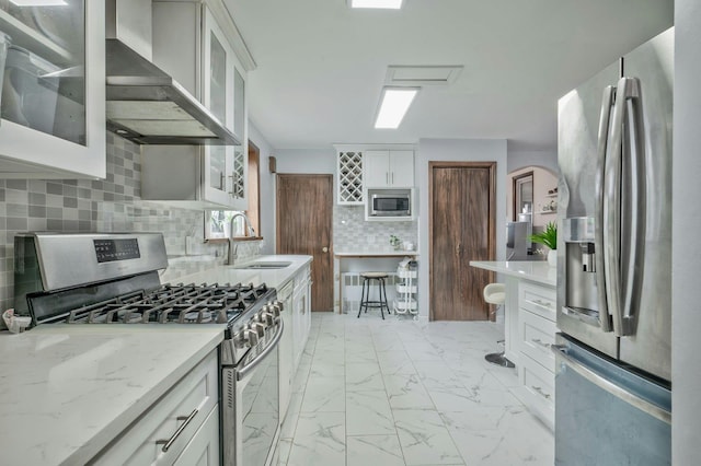 kitchen featuring a sink, white cabinets, marble finish floor, appliances with stainless steel finishes, and wall chimney exhaust hood