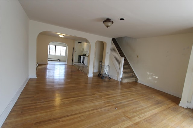 empty room featuring arched walkways, stairway, light wood finished floors, and baseboard heating