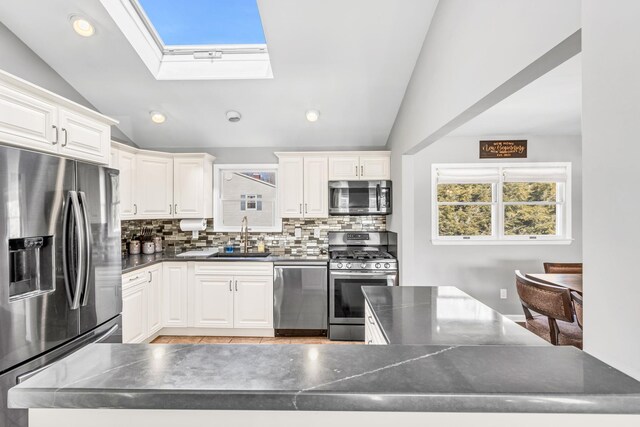 kitchen featuring stainless steel appliances, vaulted ceiling with skylight, dark countertops, and a sink