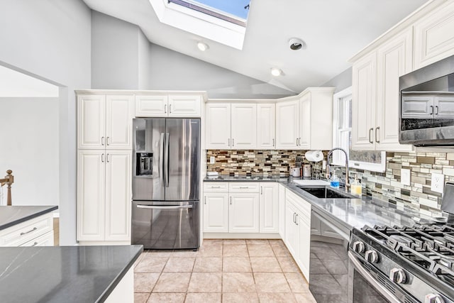 kitchen featuring dark countertops, appliances with stainless steel finishes, white cabinets, a sink, and vaulted ceiling with skylight