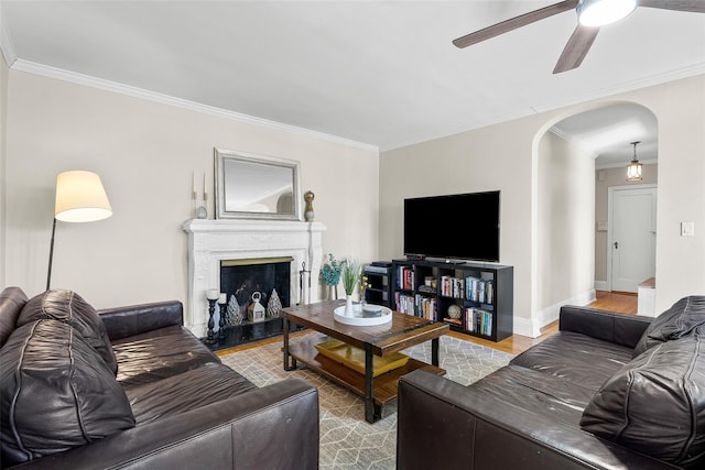 living room featuring arched walkways, crown molding, a fireplace with raised hearth, light wood-style floors, and ceiling fan