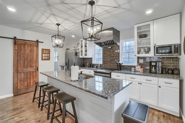 kitchen with a barn door, island range hood, stainless steel appliances, white cabinetry, and glass insert cabinets