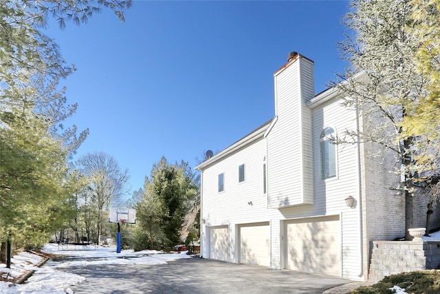 view of snowy exterior with a garage, driveway, brick siding, and a chimney