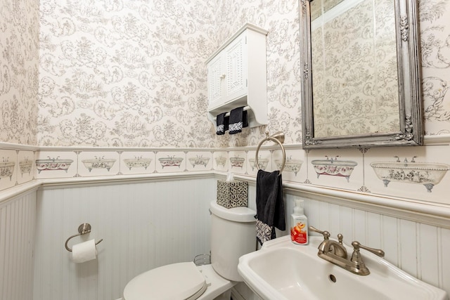 half bathroom with wainscoting, a sink, toilet, and wallpapered walls