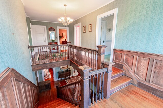 stairs with a chandelier, wood finished floors, ornamental molding, wainscoting, and wallpapered walls