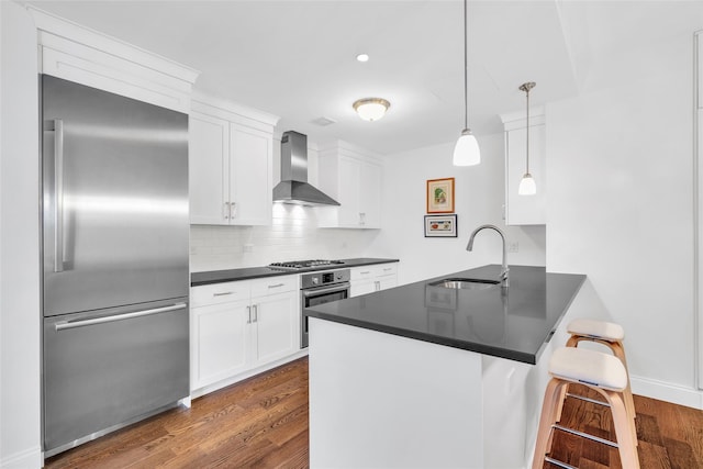 kitchen featuring a peninsula, a sink, appliances with stainless steel finishes, wall chimney range hood, and dark countertops