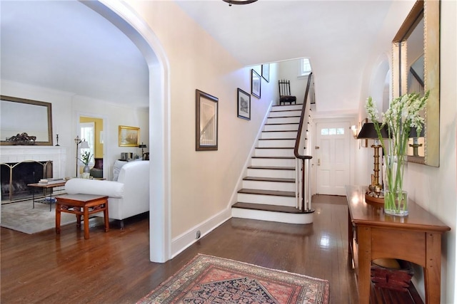 foyer entrance featuring baseboards, arched walkways, dark wood-style floors, stairway, and a fireplace