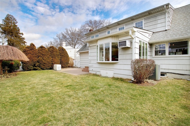 rear view of house with a shingled roof, a patio, a lawn, and a wall mounted air conditioner